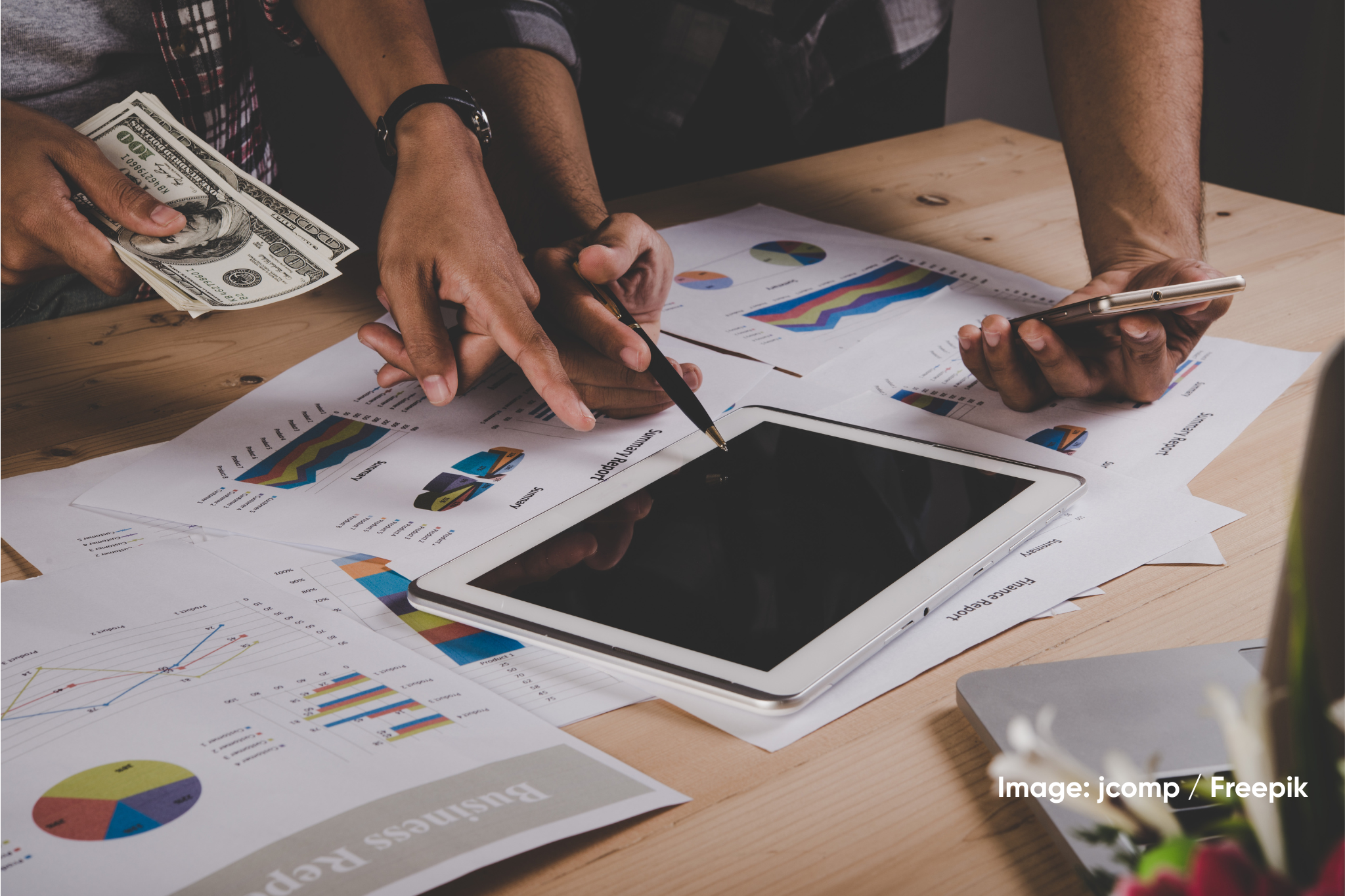 Close-up of businesspeople analyzing financial charts and graphs at a desk, with a tablet, smartphone, cash, and printed reports.